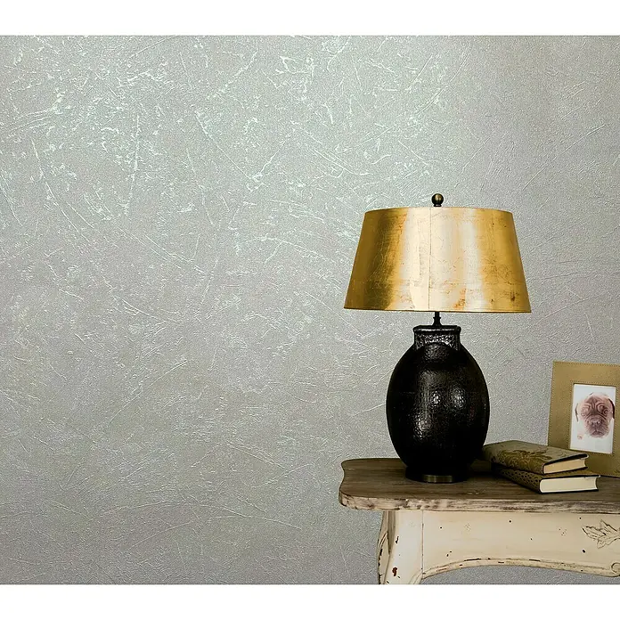 Marburg Vintage Deluxe Vliestapete - Greige-Pearl, Uni, 10,05 x 0,53 m - Nahaufnahme einer Tischlampe mit goldenem Schirm auf einem Nachttisch, die eine elegante und gemütliche Atmosphäre schafft.