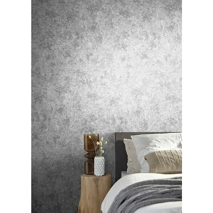 Marburg Natural Opulence Vliestapete Struktur I - Grau, Betonoptik, 10,05 x 0,7 m - Ein Schlafzimmer mit einem modernen Nachttisch, einer Lampe und einem Bett mit Kissen vor einer grauen Tapete.