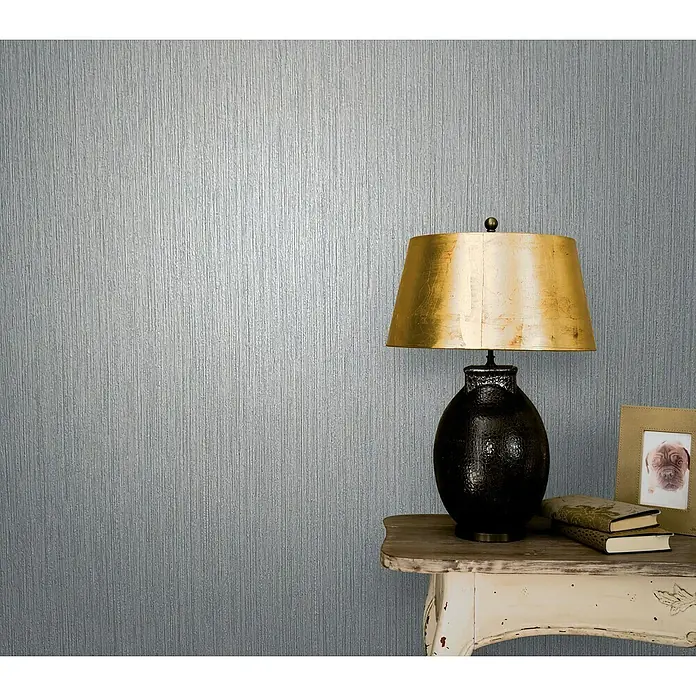 Marburg Vintage Deluxe Vliestapete - Grau, Streifen, 10,05 x 0,53 m - Nahaufnahme einer dekorativen schwarzen Lampe mit goldenem Schirm auf einem Tisch, umgeben von Büchern und einem Bilderrahmen.