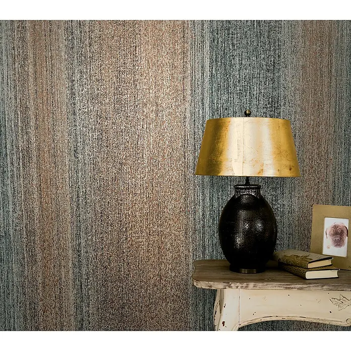 Marburg Vintage Deluxe Vliestapete - Kupfer-Anthrazit, Streifen, 10,05 x 0,53 m - Stimmungsvolle Aufnahme einer Tischlampe mit goldenem Schirm auf einem Beistelltisch, vor einer strukturierten Wand.