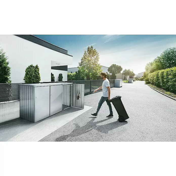 Biohort Mülltonnenbox Alex - L x B x H: 88 x 80 x 129 cm, Silber-Metallic - Silbergrauer Metall-Mülltonnenbox neben modernem Haus; Person zieht Mülltonne vorbei.