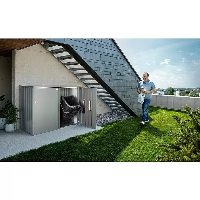 Biohort Mülltonnenbox Alex - L x B x H: 88 x 80 x 129 cm, Quarzgrau-Metallic - Silber-graues Metall-Mülltonnenbox, geöffnet mit Mülltonne, Schaufel und Besen. Ein Mann nähert sich mit Abfall.