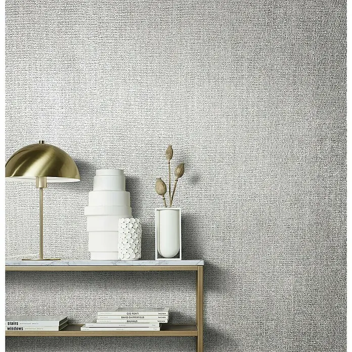Marburg Natural Opulence Vliestapete - Grau-Grün, Uni, 10,05 x 0,7 m - Nahaufnahme eines dekorierten Tisches mit einer goldenen Lampe, Vasen und Büchern, vor einer strukturierten, silbernen Wand.