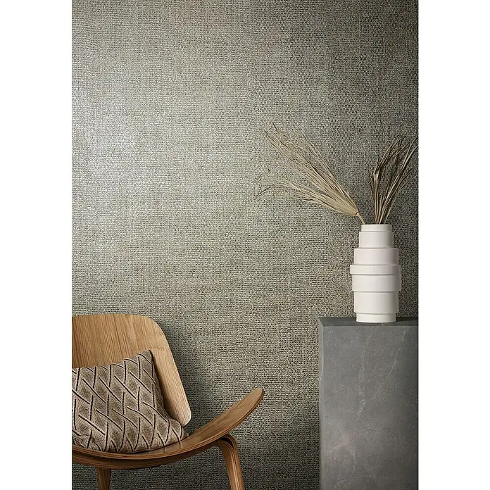 Marburg Natural Opulence Vliestapete - Beige-Braun, Uni, 10,05 x 0,7 m - Nahaufnahme eines Raumes mit strukturierten Tapeten, einem Designerstuhl mit Kissen und einer weißen Vase auf einem grauen Sockel.