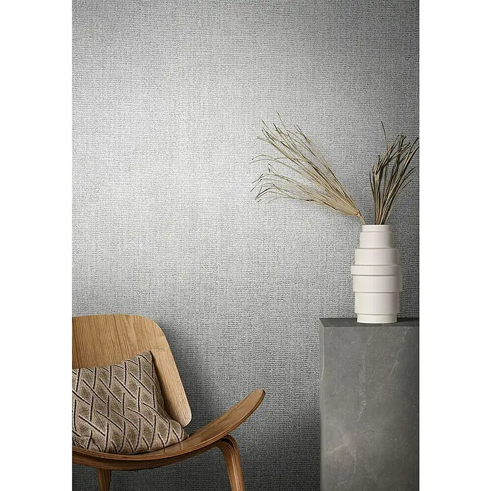 Marburg Natural Opulence Vliestapete - Grau, Uni, 10,05 x 0,7 m - Ein elegantes Interieur mit einer grauen texturierten Tapete, einem stilvollen Holzstuhl mit Kissen und einer dekorativen Vase auf einem Sockel.