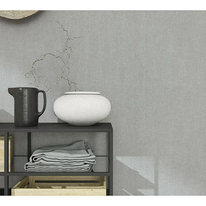 Marburg Natural Opulence Vliestapete - Grau, Uni, 10,05 x 0,7 m - Nahaufnahme eines Regals mit einer schwarzen Kanne, einer weißen Vase, gefalteten Handtüchern und Holzkisten vor einem grauen Hintergrund.
