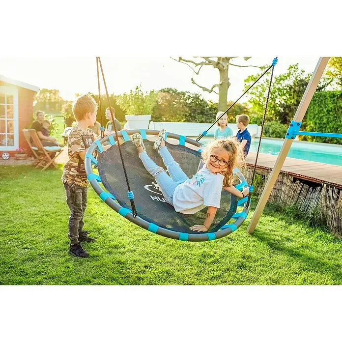 Hudora Nestschaukel Ergänzungsmodul Nest - Durchmesser: 120 cm, Stahl - Mädchen auf einer runden Hängematte im Garten, umgeben von spielenden Kindern. Die Hängematte ist blau-grau und hängt an einem Holzgestell.
