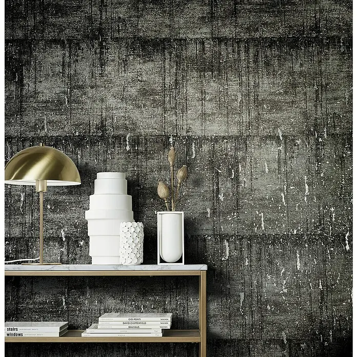 Marburg Natural Opulence Vliestapete - Schwarz-Grau, Betonoptik, 10,05 x 0,7 m - Nahaufnahme einer stilvollen Konsolentisch-Szene mit einer goldenen Lampe, weißen Vasen und Büchern vor einer strukturierten Wand.