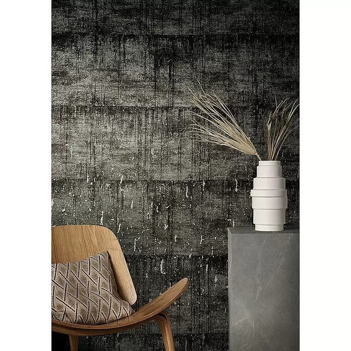 Marburg Natural Opulence Vliestapete - Schwarz-Grau, Betonoptik, 10,05 x 0,7 m - Dunkle, strukturierte Tapete mit weißen, streifigen Mustern. Davor stehen ein heller Holzstuhl mit Kissen und eine weiße Vase mit Trockenpflanzen.