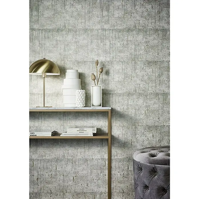 Marburg Natural Opulence Vliestapete - Helles Greige, Betonoptik, 10,05 x 0,7 m - Nahaufnahme eines stilvollen Wohnraums mit einer goldenen Lampe, weißen Vasen und einem grauen Hocker vor einer strukturierten Wand.