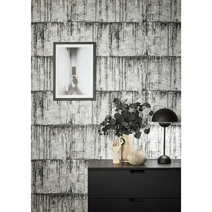 Marburg Natural Opulence Vliestapete - Schwarz-Weiß, Betonoptik, 10,05 x 0,7 m - Einrichtung mit einer schwarzen Kommode, einer Lampe und dekorativen Elementen vor einer auffälligen Wand.