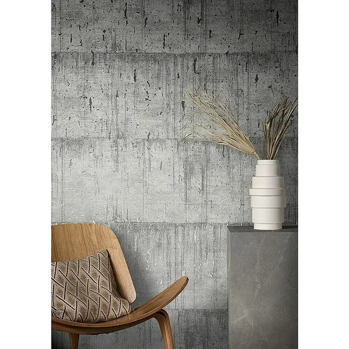 Marburg Natural Opulence Vliestapete - Grau, Betonoptik, 10,05 x 0,7 m - Einrichtungsidee mit einem hellen Holzstuhl mit Kissen, einer weißen Vase mit getrockneten Gräsern vor einer grauen Wand.