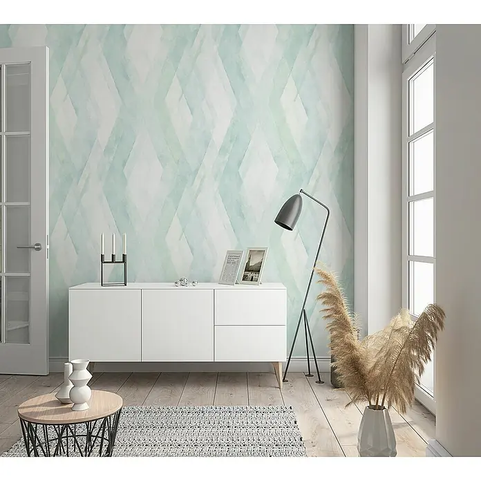 Marburg Shades Vliestapete - Grün-Weiß, Grafisch, 10,05 x 0,53 m - Das Bild zeigt ein stilvolles Wohnzimmer mit einer weißen Kommode, dekoriert mit Kerzenhaltern und Fotorahmen, vor einer gemusterten Tapete.