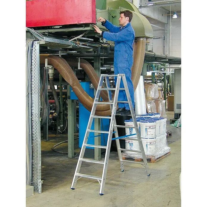 Krause Stabilo Doppelleiter - Anzahl Tritte: 16 Stk., Arbeitshöhe: 3,4 m, Aluminium - Eine silberne Krause-Leiter steht in einer Fabrik, auf der ein Mann in blauer Arbeitskleidung an einer Maschine arbeitet.
