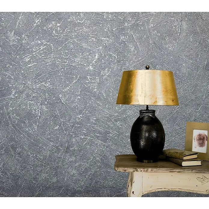 Marburg Vintage Deluxe Vliestapete - Grau-Silber, Uni, 10,05 x 0,53 m - Elegante Tischleuchte mit goldenem Schirm und dunklem Sockel auf einem Beistelltisch, Bücher und Bilderrahmen daneben.