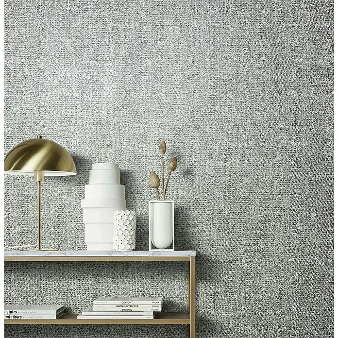 Marburg Natural Opulence Vliestapete - Grau-Silber, Uni, 10,05 x 0,7 m - Ein stilvolles Interieur mit einer eleganten Konsole, dekoriert mit Vasen, einer Lampe und Büchern vor einer strukturierten Tapete.