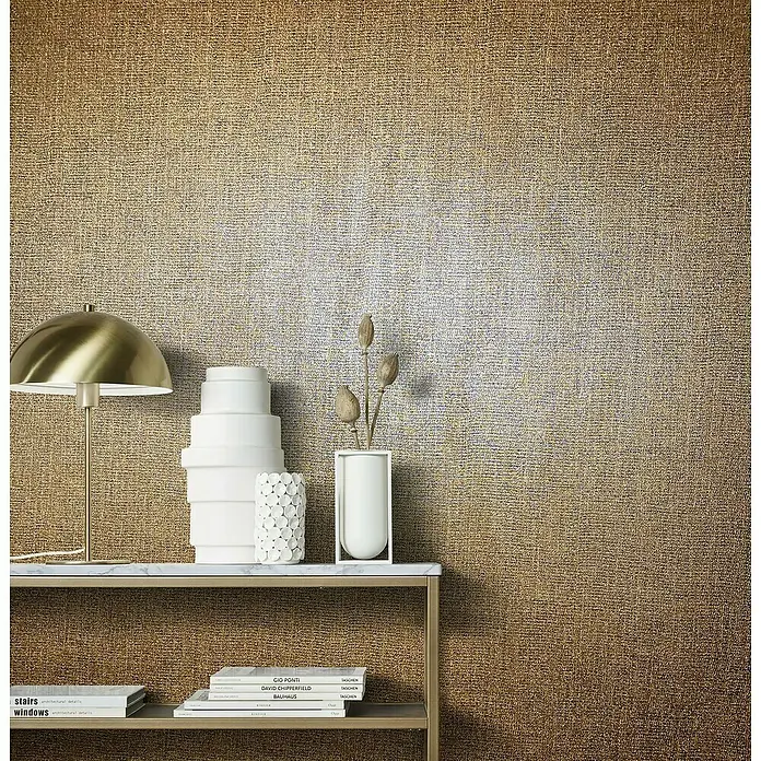 Marburg Natural Opulence Vliestapete - Kupfer, Uni, 10,05 x 0,7 m - Elegante Tischleuchte mit goldenem Schirm auf einem Regal mit dekorativen Büchern und Vasen vor einer strukturierten Wand.