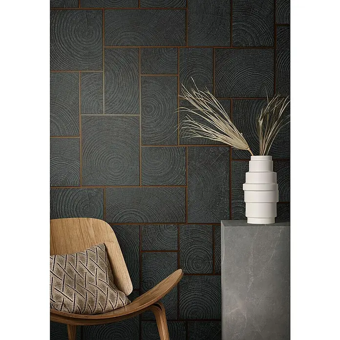 Marburg Natural Opulence Vliestapete - Anthrazit, Holzoptik, 10,05 x 0,7 m - Nahaufnahme einer Wand mit quadratischen, dunkelgrauen Fliesen im Holzdesign, einem weißen Dekorationselement und einem Stuhl mit Kissen.