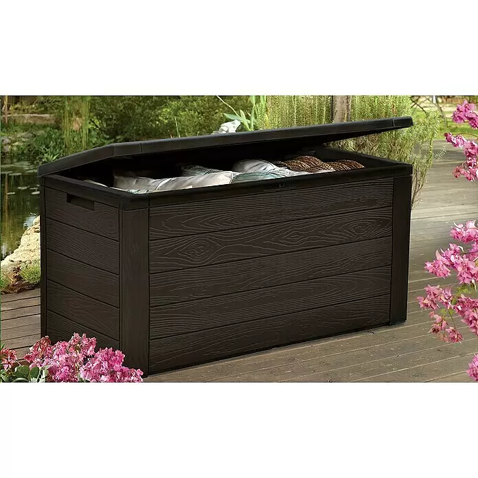 Jelenia Plast Garten-Aufbewahrungsbox Woody - 119 x 45 x 57 cm, Kunststoff, Anthrazit - Dunkelbraune Outdoor-Aufbewahrungsbox in Holzoptik, Deckel offen, auf einer Terrasse, lagert Kissen.