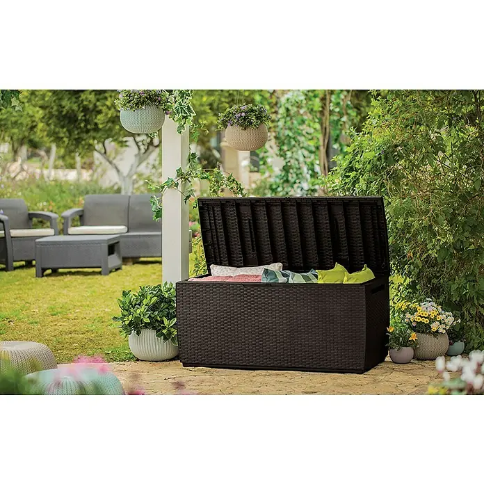 Keter Baúl para jardín Capri - 54 x 123 x 57 cm, Marrón - Caja de jardín de ratán marrón con tapa abierta llena de cojines, en un patio. Muebles de jardín y plantas al fondo.