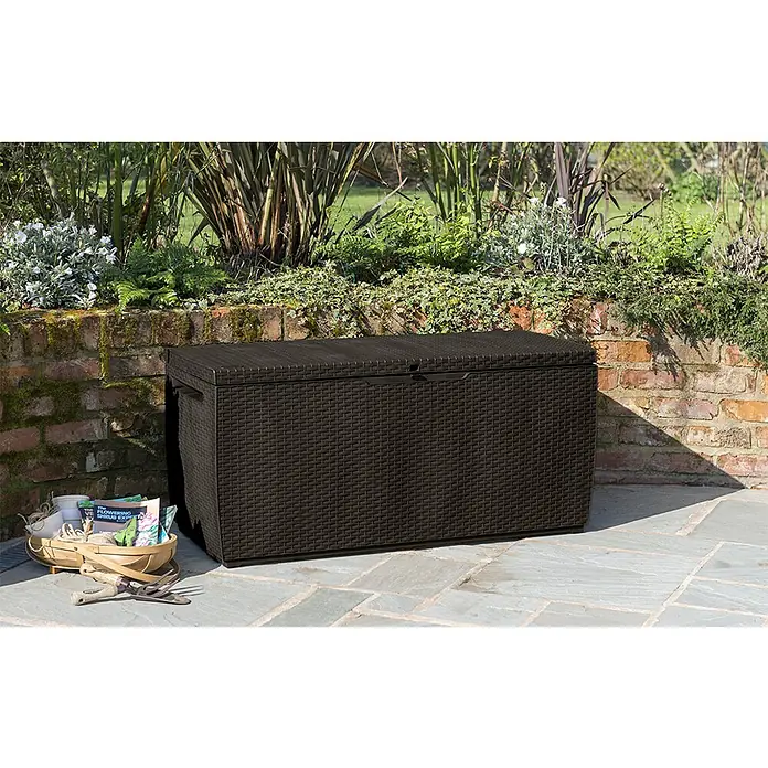 Keter Baúl para jardín Capri - 54 x 123 x 57 cm, Marrón - Una caja de jardín de ratán marrón se encuentra en un patio, ideal para guardar herramientas de jardín y cojines.