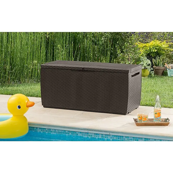 Keter Baúl para jardín Capri - 54 x 123 x 57 cm, Marrón - Una caja de almacenamiento Keter marrón está colocada junto a una piscina, junto con un pato inflable y una botella de jugo de naranja.