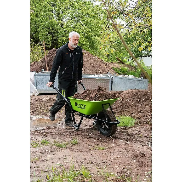 Zipper Akku-Schubkarre ZI-EWB500LI - 500 W, Material Wanne: Stahl, Traglast: 150 kg - Ein Mann schiebt eine grüne Zipper ZI-EWB500Li Schubkarre voller Erde durch einen Garten mit Erdhügeln.