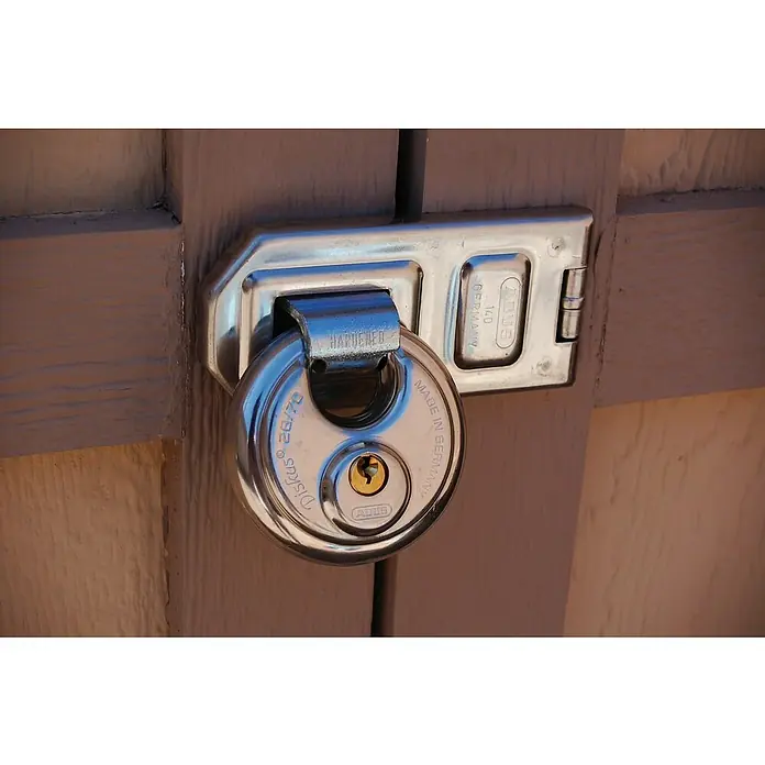 Abus Diskus-Überfalle 140 - Maße Überfalle: 120 x 56 mm, Durchmesser Öse: 12,2 mm, Edelstahl - Nahaufnahme eines Abus Diskus 26/70 Vorhängeschlosses, das an einem silberfarbenen Riegel an einer Holztür befestigt ist, der Sicherheit und Schutz bietet.