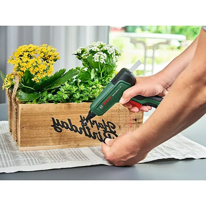 Bosch Power for All 18V Akku-Heißklebepistole AdvancedGlue 18V - 18 V, Ohne Akku, Temperaturbereich: 130 °C - 200 °C - Eine Person verwendet eine Bosch AdvancedGlue 18V Klebepistole, um schwarze Buchstaben auf einer Holzkiste mit Blumen zu befestigen.