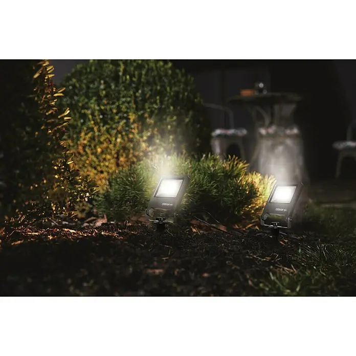 Ledvance LED-Gartenstrahler Endura Floodspike - 20 W, 1.700 lm, 3.000 K, 4,4 x 13,3 x 32,3 cm, IP65 - Gartenbeleuchtung mit zwei Strahlern der Marke OSRAM, die Büsche und einen Tisch im Hintergrund beleuchten.\n