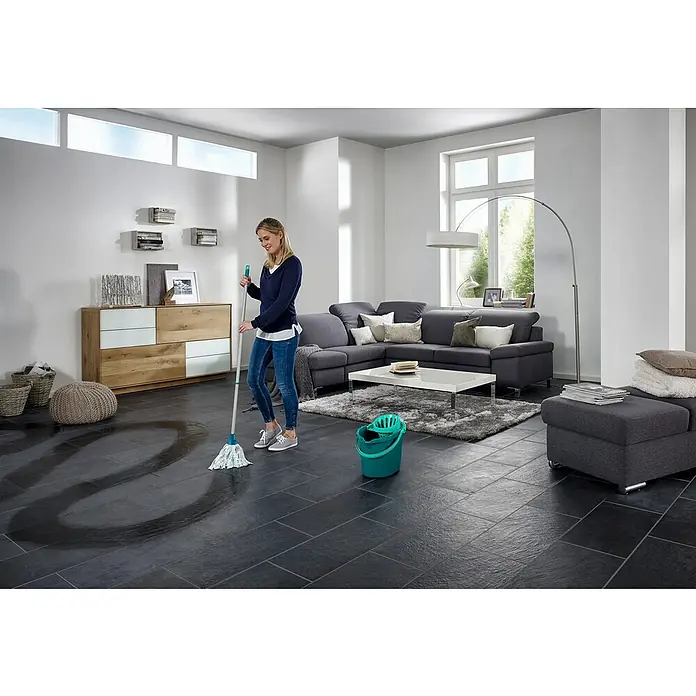 Leifheit Set de limpieza Classic - 12 l, Azul - Mujer limpia el suelo de baldosas oscuras con una fregona, el cubo está al lado. El moderno salón está amueblado con luminosidad.