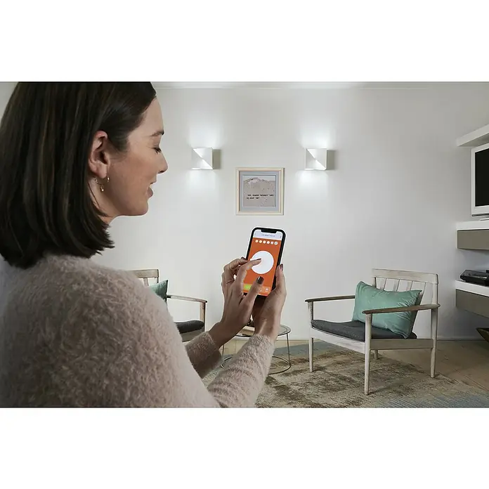 Ledvance LED-Wandleuchte - 23 W, L x B x H: 30 x 15 x 10,5 cm, Weiß, Mehrfarbig - Frau bedient das Smartphone zur Steuerung der intelligenten Beleuchtung, die an der Wand montiert ist, für eine einfache und bequeme Bedienung.