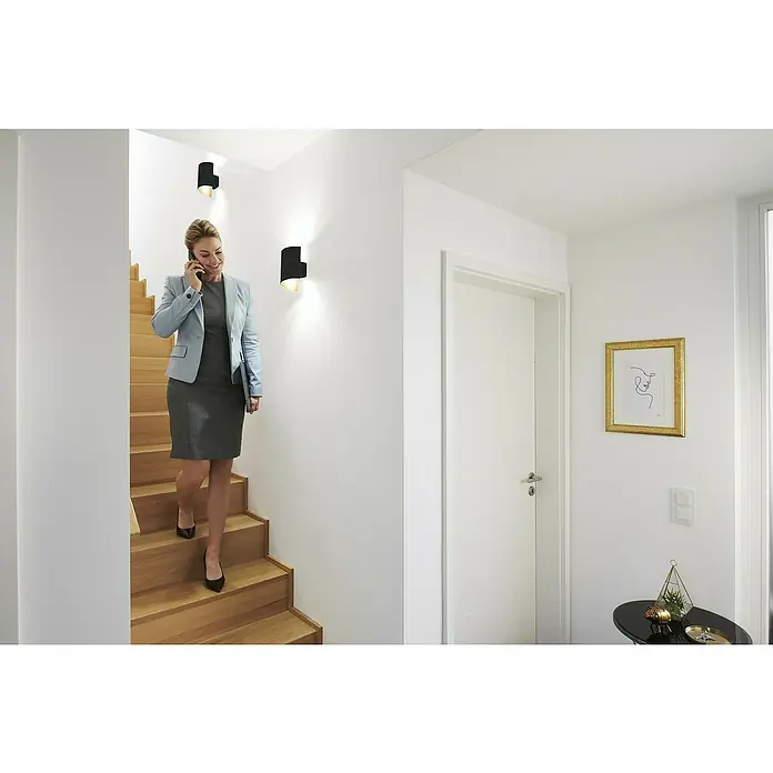 Ledvance Smart+ WiFi LED-Wandleuchte - 12 W, L x B x H: 12,7 x 9,5 x 23 cm, Schwarz, Warmweiß - Eine Frau geht die Treppe hinunter, beleuchtet von modernen Wandleuchten, die ein warmes Licht spenden. Elegante Beleuchtung für ein stilvolles Zuhause.