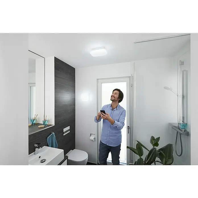 Ledvance LED-Wandleuchte - 12 W, L x B x H: 20 x 20 x 9,4 cm, Weiß, Mehrfarbig - Ein Mann mit einem Smartphone in einem Badezimmer mit einer quadratischen Deckenleuchte. Das Licht sorgt für eine angenehme Atmosphäre.
