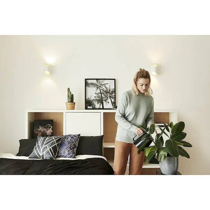 Ledvance LED-Wandleuchte12 W, L x B x H: 12,7 x 9,5 x 20 cm, Weiß, Mehrfarbig - Eine junge Frau gießt eine Zimmerpflanze mit einer eleganten, schwarzen Gießkanne in einem modernen, hellen Wohnraum.