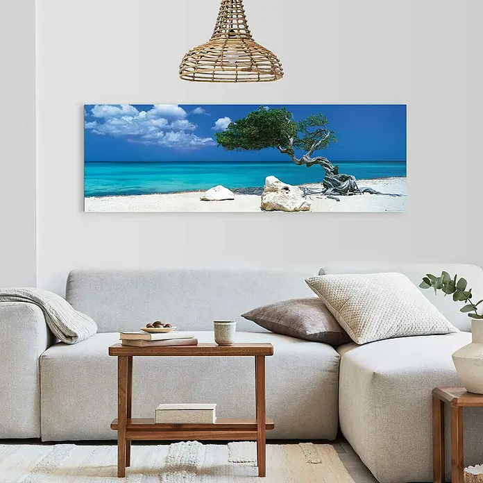Reinders Decopanel - Divi Divi Tree, B x H: 156 x 52 cm - Ein modernes Wohnzimmer mit einem hellen Sofa, einem Holztisch und einem Landschaftsbild einer tropischen Szene mit einem markanten Baum.