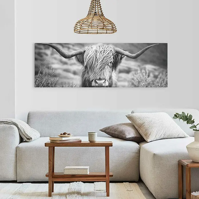 Reinders Decopanel - Highlander Landscape, B x H: 156 x 52 cm - Wohnzimmer mit einem grauen Sofa, einem Holztisch und einem Schwarz-Weiß-Bild eines Highland-Rinds an der Wand. Eine Rattalampe hängt von der Decke.