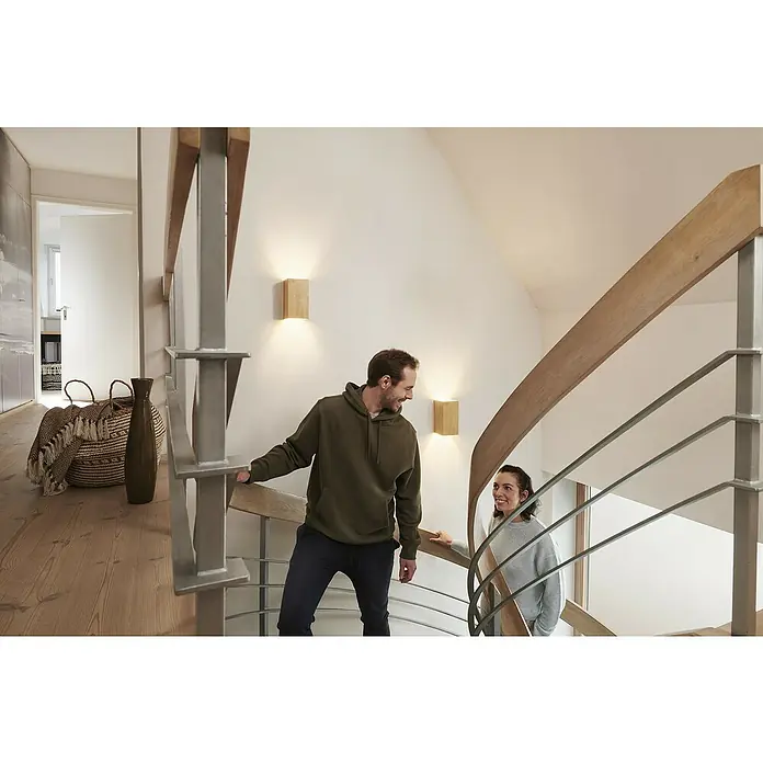 Ledvance Smart+ WiFi LED-Wandleuchte - 12 W, L x B x H: 11 x 11 x 21 cm, Gelb, Warmweiß - Ein Paar steigt eine Treppe hinauf, beleuchtet durch moderne Holz-Wandleuchten, die warmes Licht spenden und eine angenehme Atmosphäre schaffen.