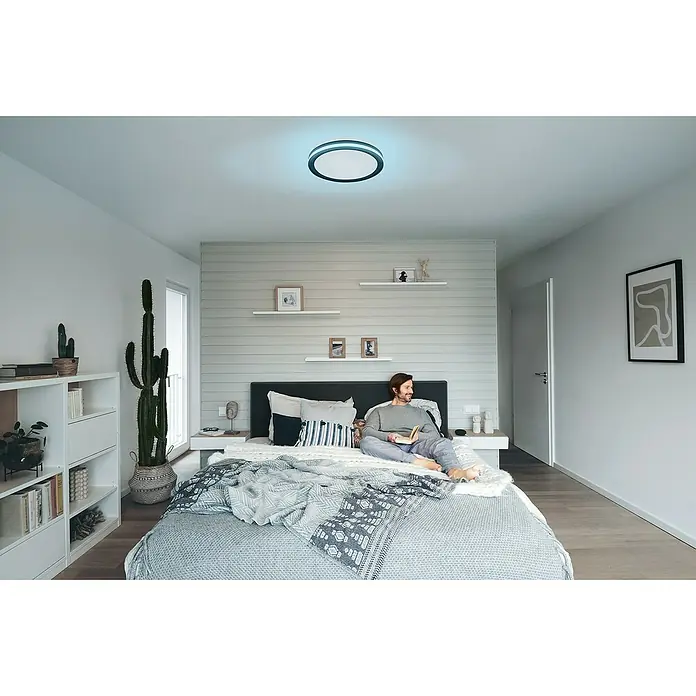 Ledvance LED-Deckenleuchte rund - 28 W, Ø x H: 46 x 9,5 cm, Schwarz, Mehrfarbig - Deckenleuchte mit farbigem Rand, die ein Zimmer mit hellen Wänden und einem Mann im Bett beleuchtet, der ein Buch liest.