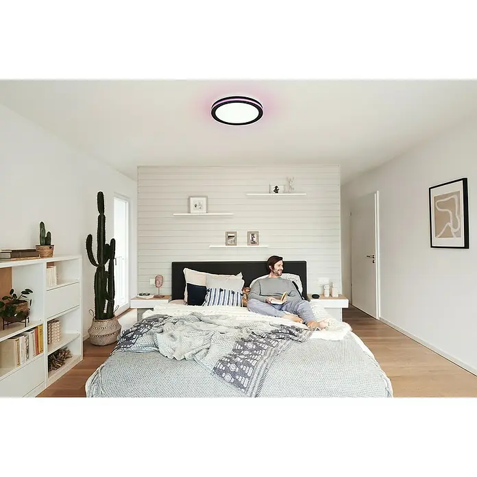Ledvance LED-Deckenleuchte rund - 28 W, Ø x H: 46 x 9,5 cm, Schwarz, Mehrfarbig - Blick in ein Schlafzimmer mit einer modernen runden Deckenleuchte mit farbigen Akzenten, die über einem Mann im Bett hängt.