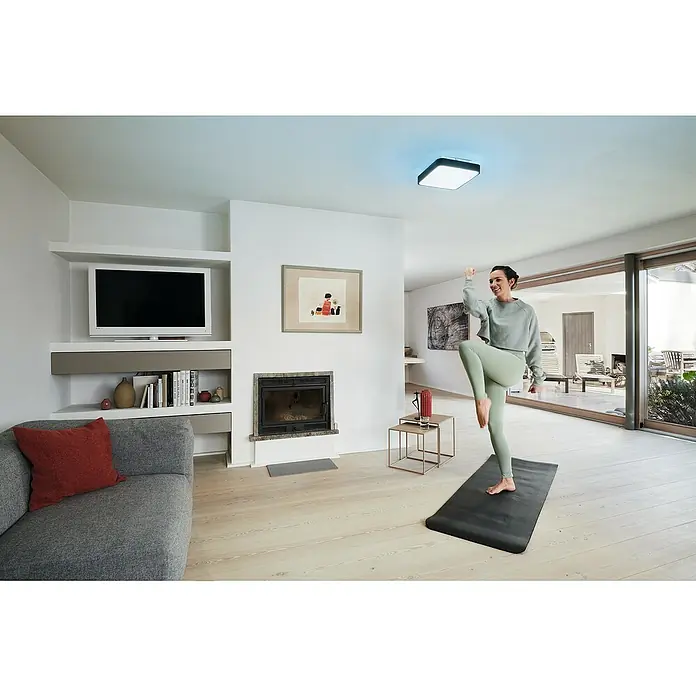 Ledvance LED-Deckenleuchte - 28 W, L x B x H: 35 x 35 x 9 cm, Schwarz, RGBW - Helle Wohnung mit einer schlanken Deckenleuchte, die modernes Design und Funktionalität vereint, während eine Frau Yoga praktiziert.