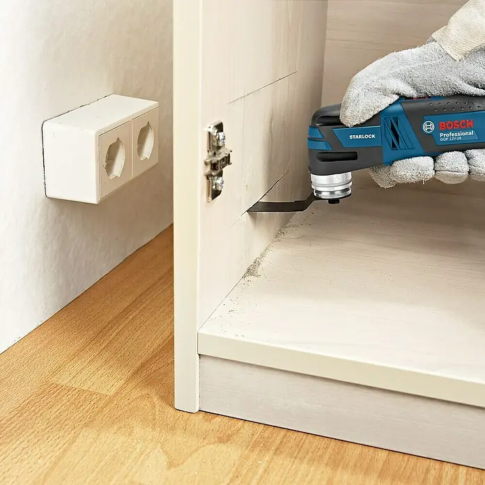 Bosch Professional Maschinen-Set 5-Tool-Kit GSR+GOP+GDR+GSA+GLI - 12 V, 3 Akkus, 2 Ah, 10 -tlg. - Bosch Professional GOP 12V-28 oszillierendes Multifunktionswerkzeug im Einsatz beim Schneiden eines Schranks in der Nähe einer Steckdose.