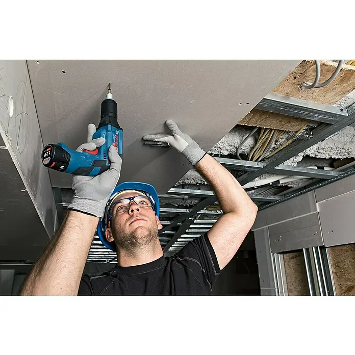 Bosch Professional Akku-Trockenbauschrauber GTB 12V-11 - 12 V, Ohne Akku, Ohne Koffer - Ein Bauarbeiter befestigt eine Gipskartondecke mit einem blauen Bosch 12V Akkuschrauber, Schutzbrille und Helm für Sicherheit.