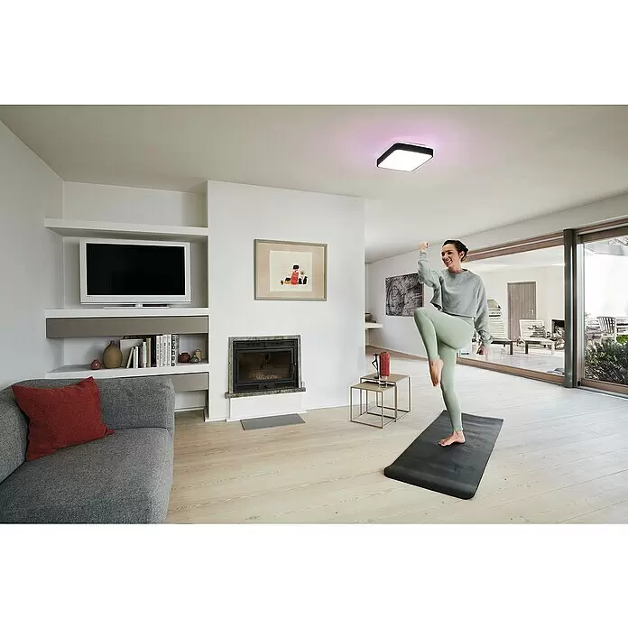 Ledvance LED-Deckenleuchte - 28 W, L x B x H: 35 x 35 x 9 cm, Schwarz, RGBW - Quadratische schwarze Smart-Deckenleuchte an weißer Decke, die eine Frau beim Sport in einem modernen Wohnzimmer beleuchtet.