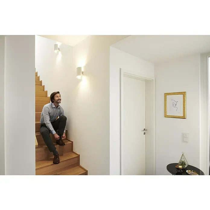 Ledvance Smart+ WiFi LED-Wandleuchte - 12 W, L x B x H: 12,7 x 9,5 x 23 cm, Weiß, Warmweiß - Ein Mann sitzt auf einer Holztreppe unter zwei Wandleuchten und bindet seine Schnürsenkel. Stilvolle Beleuchtung für Zuhause.