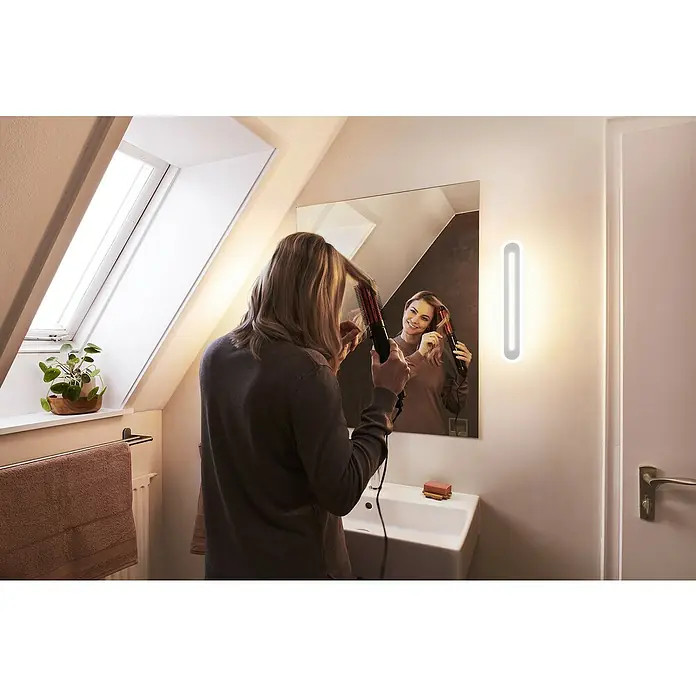 Ledvance Smart+ WiFi LED-Wandleuchte - 13 W, L x B x H: 30 x 7 x 5 cm, Weiß, Warmweiß - Eine Frau stylt ihre Haare vor einem beleuchteten Spiegel im Badezimmer. Der Fokus liegt auf der Badezimmerbeleuchtung.