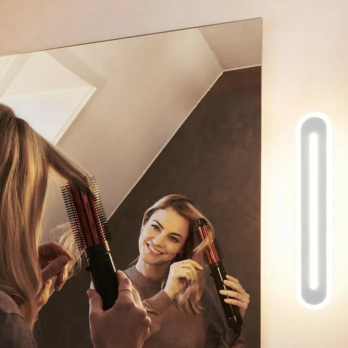 Ledvance Smart+ WiFi LED-Wandleuchte - 13 W, L x B x H: 30 x 7 x 5 cm, Weiß, Warmweiß - Frau stylt ihr Haar mit einer rotierenden Heißluftbürste vor einem Spiegel. Eine Lampe wirft ein sanftes Licht auf die Szene.