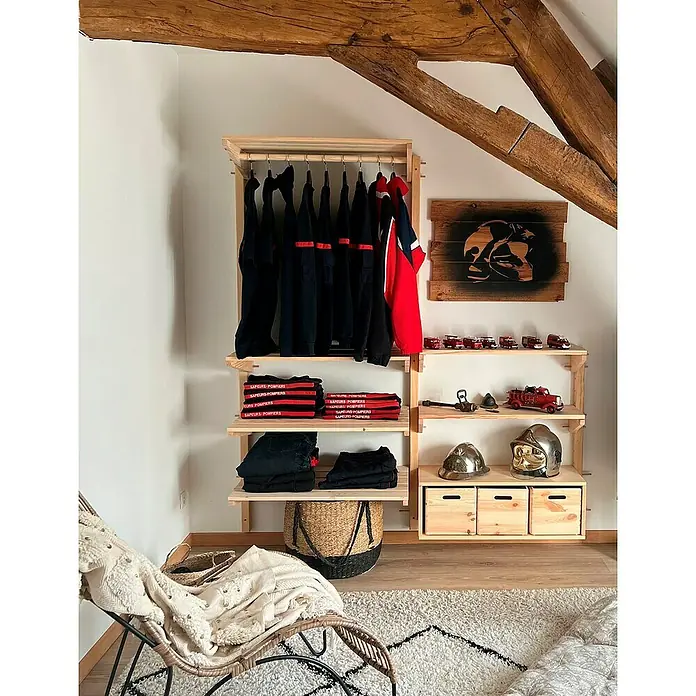 Astigarraga Wally Estante de madera - L x An x Al: 46 x 80 x 13,7 cm, Natural, Madera de pino, Carga límite balda: 100 kg - Un armario de madera clara con ropa de bombero, cascos y coches de juguete, contra un techo inclinado.