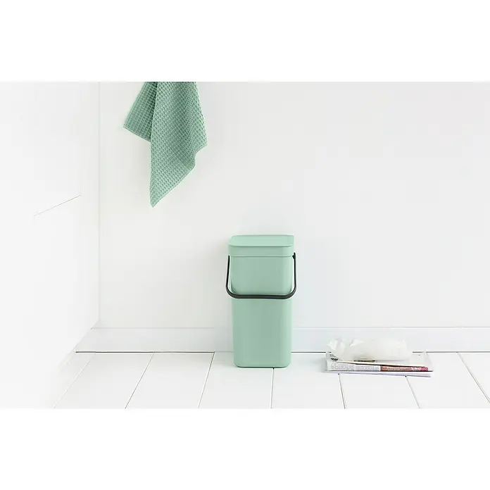 Brabantia Cubo de basura empotrado Sort & Go - 12 l, Verde, Plástico - Cubo de basura de plástico color menta con tapa y asa negra, al lado de un periódico y un pañuelo delante de una pared blanca.