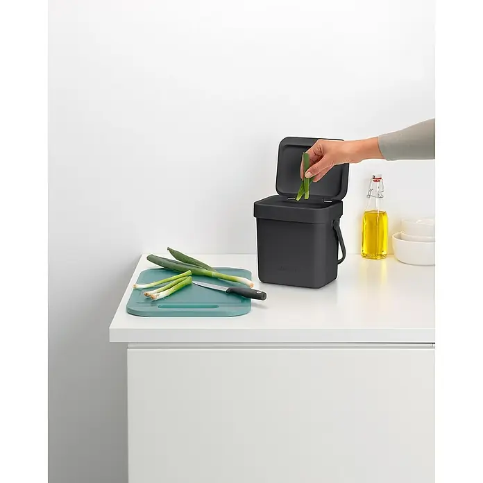 Brabantia Cubo de basura empotrado Sort & Go - 3 l, Plástico, Negro - Un contenedor de compost gris con tapa abierta está sobre una encimera de cocina mientras una mano tira residuos verdes.